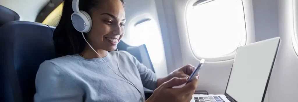 A woman on an airplane listening to music with her wireless noise-cancelling headphones, demonstrating essential travel items for a comfortable journey.