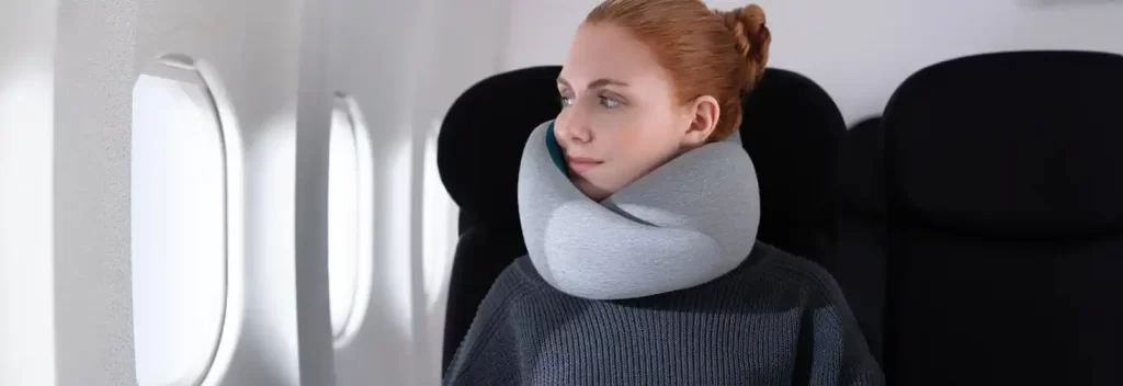 A woman on an airplane resting comfortably with a neck pillow, illustrating one of the essential travel items for a relaxing flight.