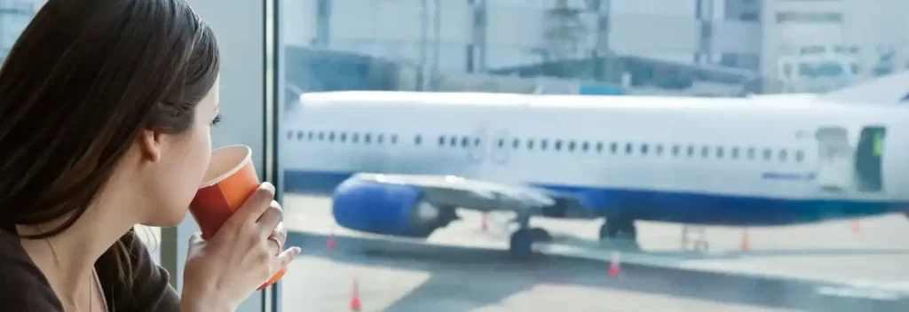 A woman at her airport gate, drinking from a cup while looking at the plane through the window, showcasing a moment of relaxation before boarding.