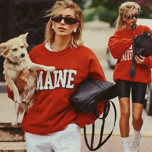 Hailey Bieber effortlessly rocks the perfect classic comfort soccer mom outfit. She is dressed in an oversize red sweatshirt, paired with black bike shorts and white sneakers. She accessorizes with stylish black sunglasses and a large black handbag. Her look is casual yet stylish, perfect for a comfy and practical day at the soccer field. 