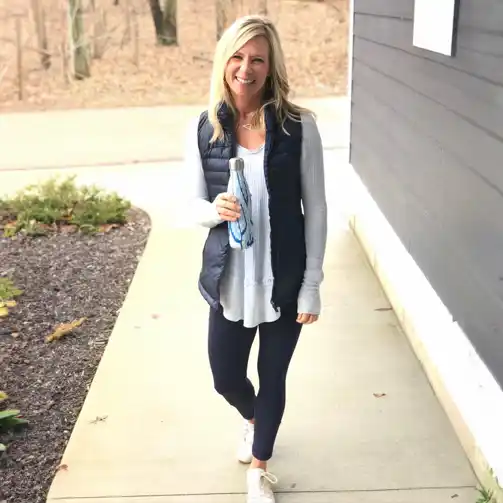 This soccer mom showcases a perfect layered look for game day. She is wearing a light gray long-sleeve top underneath a navy puffer vest, paired with black leggings and white sneakers. She holds a reusable water bottle, completing her practical and stylish outfit. This ensemble is ideal for staying warm and comfortable during cooler weather, making it perfect for a day spent on the sidelines.