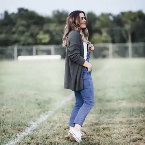 This soccer mom perfectly exemplifies the layered up look for the field. She is dressed in a comfortable ensemble featuring blue denim overalls, a white t-shirt, and a cozy dark green cardigan. Her casual yet stylish outfit is completed with white sneakers, making it ideal for staying warm and comfortable during unpredictable weather. This look is practical and versatile, ensuring that she is prepared for any sideline situation.