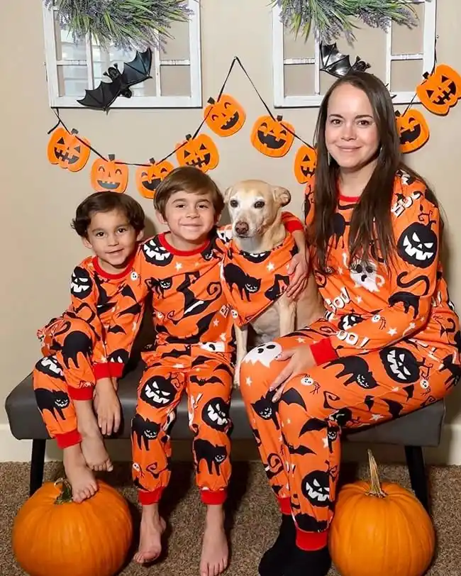 Family dressed in matching family Halloween pajamas with a pumpkin and black cat pattern, posing with their dog in front of Halloween decorations for a festive look.