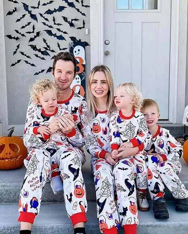 Family sitting on the porch wearing matching family Halloween pajamas with colorful Halloween designs, including pumpkins, ghosts, and spider webs, with Halloween decorations in the background.