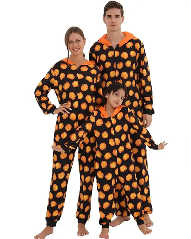 Family wearing matching pumpkin-themed onesie pajamas for Halloween, featuring a black background with vibrant orange pumpkin prints, posing together and smiling.