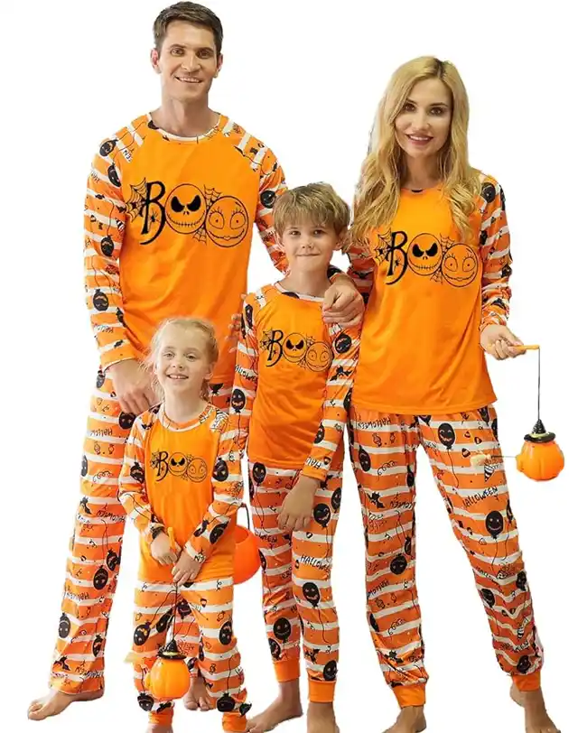 Family dressed in matching Halloween pajamas featuring an orange and black 'Boo!' design with smiling jack-o'-lanterns and stripe patterns, holding pumpkin lanterns and smiling together.