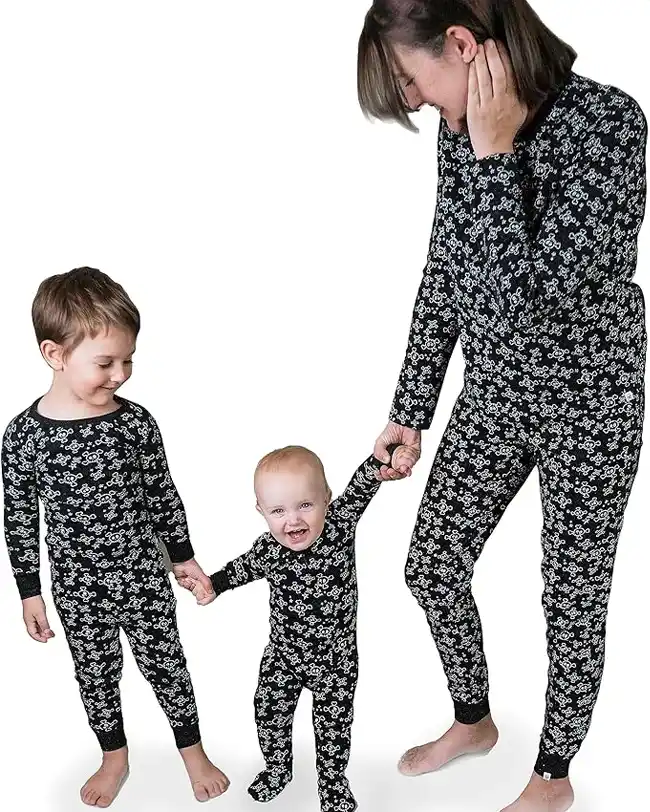 Mom and two kids dressed in matching Halloween pajamas featuring a black background with white skull and bone patterns, holding hands and smiling together.