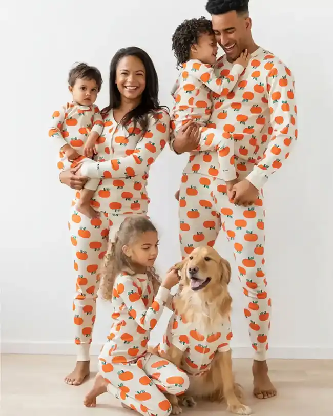 Family dressed in matching Halloween pajamas featuring an orange pumpkin pattern, including mom, dad, two kids, and a dog, all smiling and posing together.