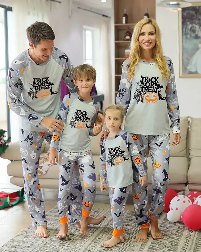 Family dressed in matching gray Halloween pajamas with 'Trick or Treat' text and various Halloween motifs, including mom, dad, two children, all smiling and standing together in a cozy living room.