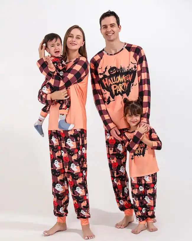 Family dressed in matching Halloween pajamas featuring orange and black checkered tops with 'Halloween Party' text and various Halloween motifs, including mom holding a baby, dad, and two children, all smiling together.