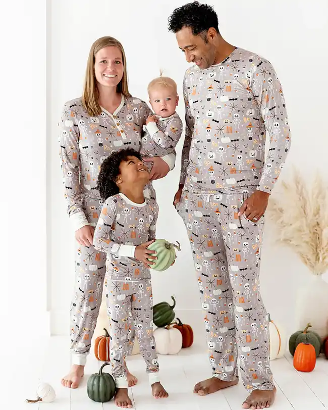 Family of four dressed in matching Halloween pajamas with a festive design featuring skulls, bats, and pumpkins. The pajamas have a light gray background with playful Halloween elements in orange, black, and white. The family is standing together, holding pumpkins, and smiling.