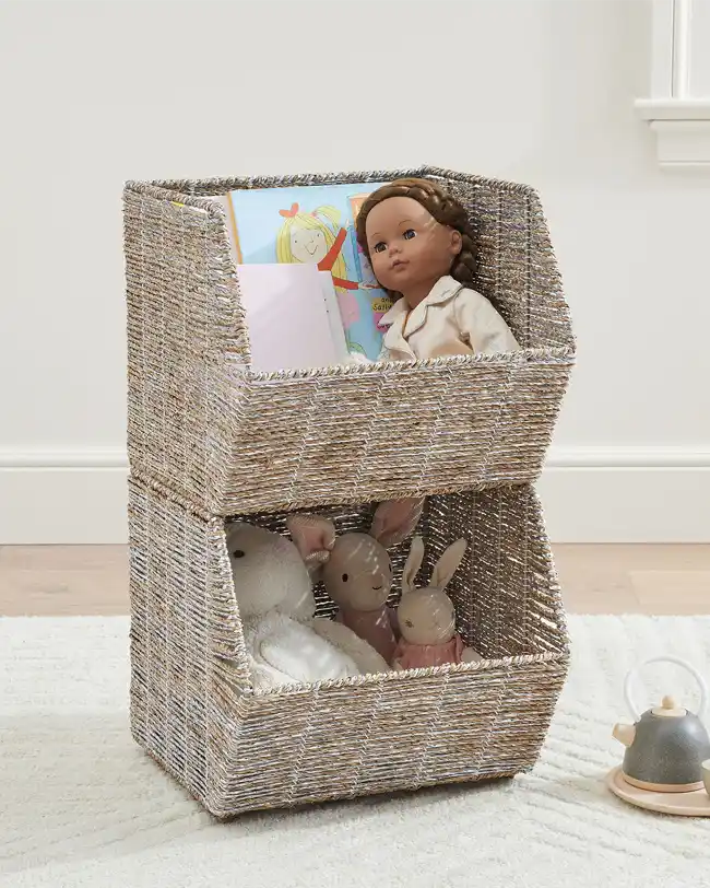 Two woven stacking toy baskets, one on top of the other, filled with dolls, stuffed animals, and books, providing a natural and stylish toy storage solution.