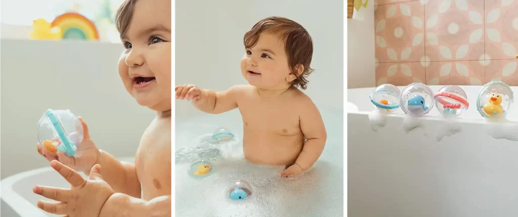 A happy baby playing in the bath with Munchkin Float and Play Bubbles baby bath toys. The baby is seen holding one of the clear bubbles with a blue and orange sea creature inside, while other bubbles float in the bathwater and rest on the bathtub edge.