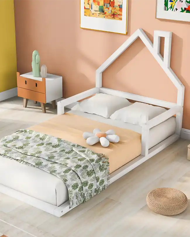 A cozy toddler floor bed designed in a Montessori style, featuring a white wooden frame with a charming house-shaped headboard. The bed is placed in a modern, pastel-colored bedroom, perfect for promoting independence and self-reliance in young children.