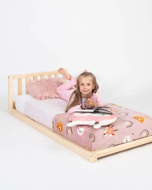 A young girl lying on a toddler floor bed designed in a Montessori style, made from natural wood. The bed features a low profile with a simple, open frame, encouraging independence. The girl is comfortably reading a book, demonstrating the cozy and inviting nature of this Montessori bed.