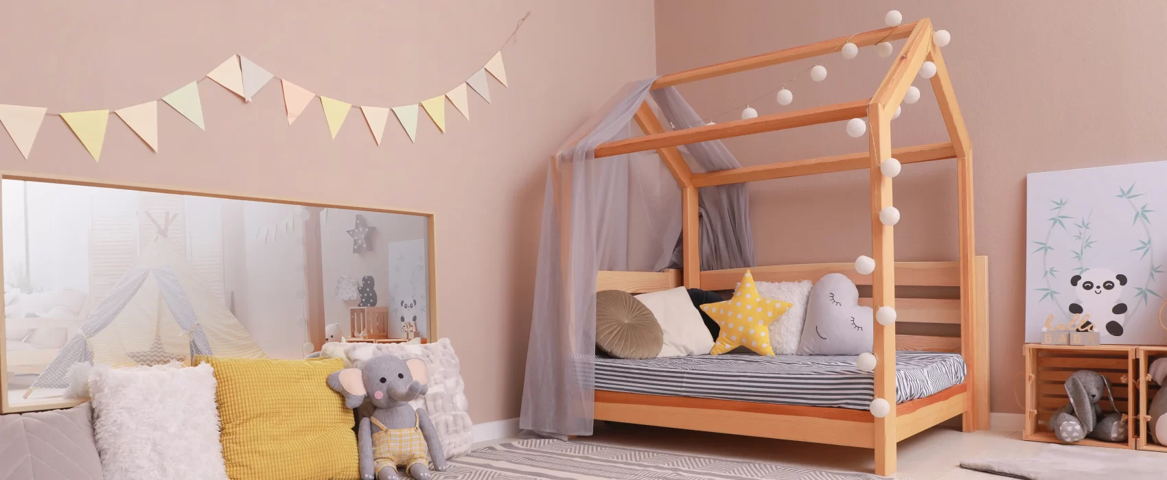 A cozy Montessori floor bed featuring a wooden house-shaped frame adorned with string lights. The toddler floor bed has a low profile, promoting independence for young children. The bed is set in a warm and inviting room with pastel decor, including decorative pillows, a soft rug, and a playful bunting banner. The setting creates a perfect environment for rest and imaginative play, fostering a sense of ownership and comfort.