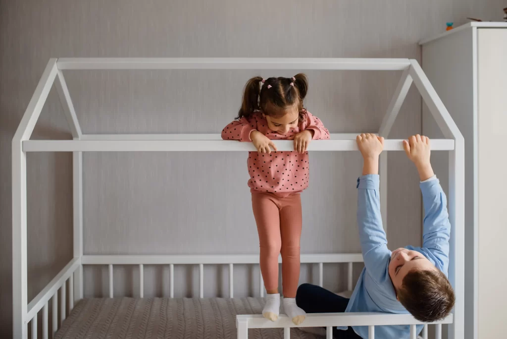 Two young children playing on a white Montessori floor bed with a house-shaped frame. The toddler floor bed has a low profile, encouraging independence and imaginative play. The children are enjoying themselves, demonstrating the playful and engaging design of the Montessori bed, perfect for fostering a sense of ownership and creativity in their sleep space.