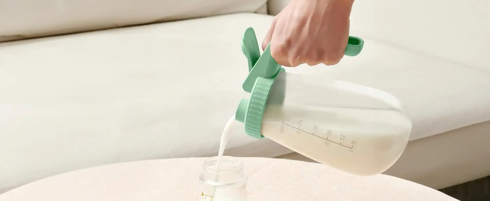 A hand holding a large BPA-free pitcher while pouring breast milk into a bottle, demonstrating the pitcher method for efficient breast milk storage.