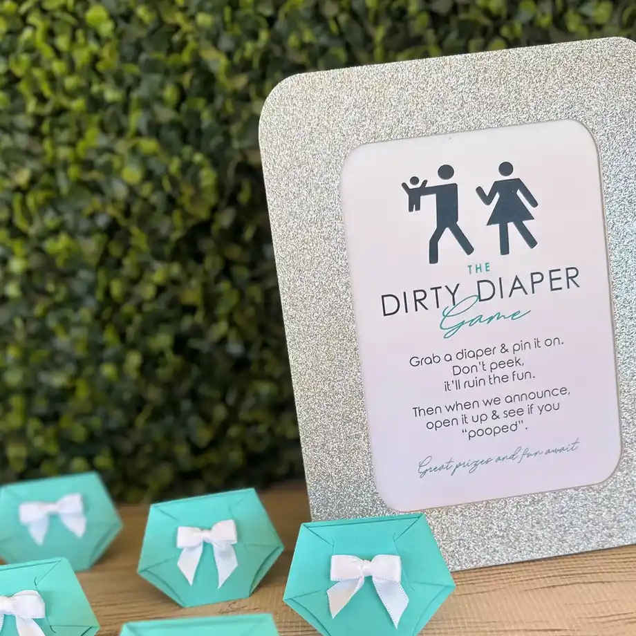 A glittery display board for the Dirty Diaper Game, surrounded by turquoise mini diaper props for a festive baby shower game.