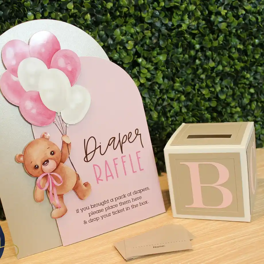 A pink diaper raffle sign featuring a teddy bear with balloons and a matching ticket box, designed for a baby shower game.