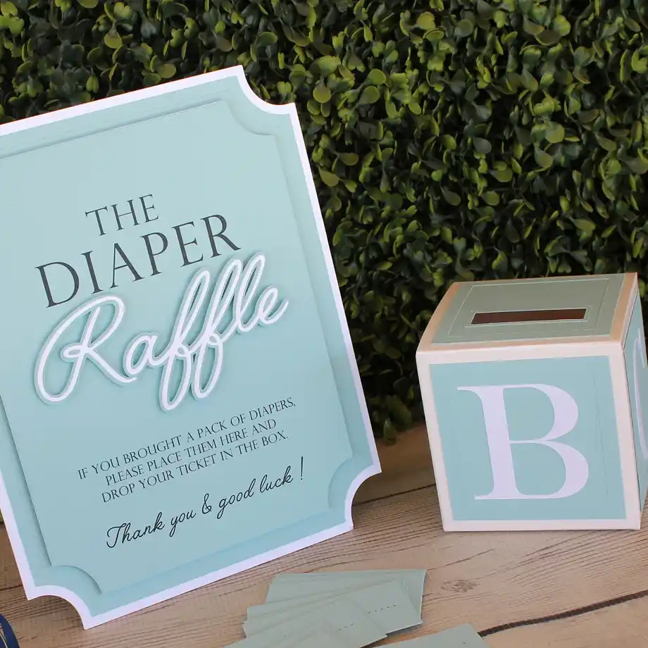 A sophisticated blue diaper raffle sign with a matching ticket box, set against a leafy backdrop for a baby shower game.