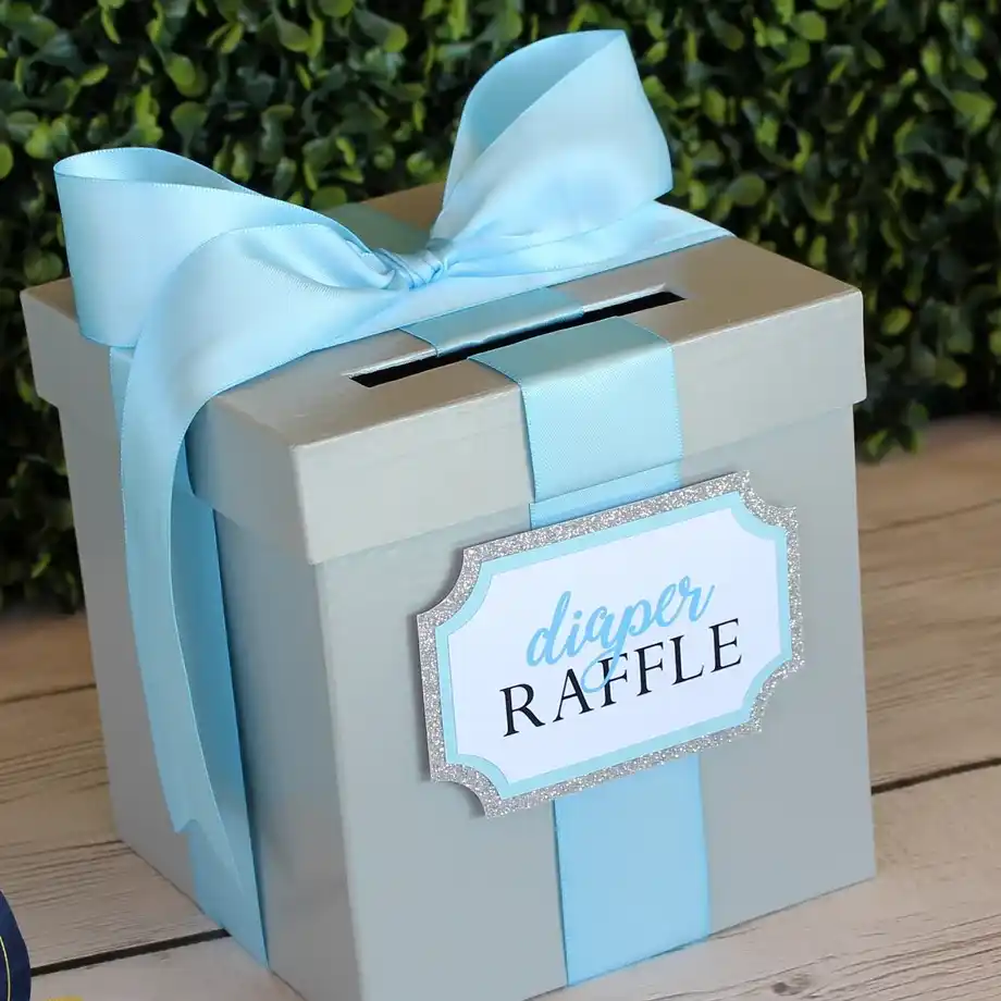 A blue gift box-style diaper raffle collection box with a ribbon and decorative label, ideal for a baby shower game.