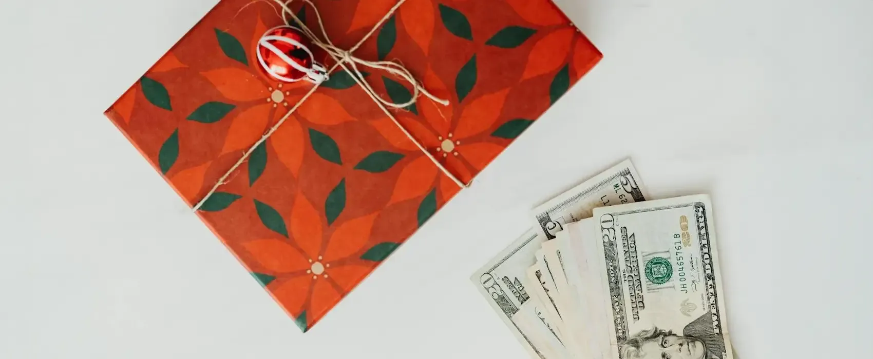 A wrapped red holiday gift box with green floral patterns and a small ornament, paired with dollar bills arranged neatly on a white background, showcasing fun ways to gift money.