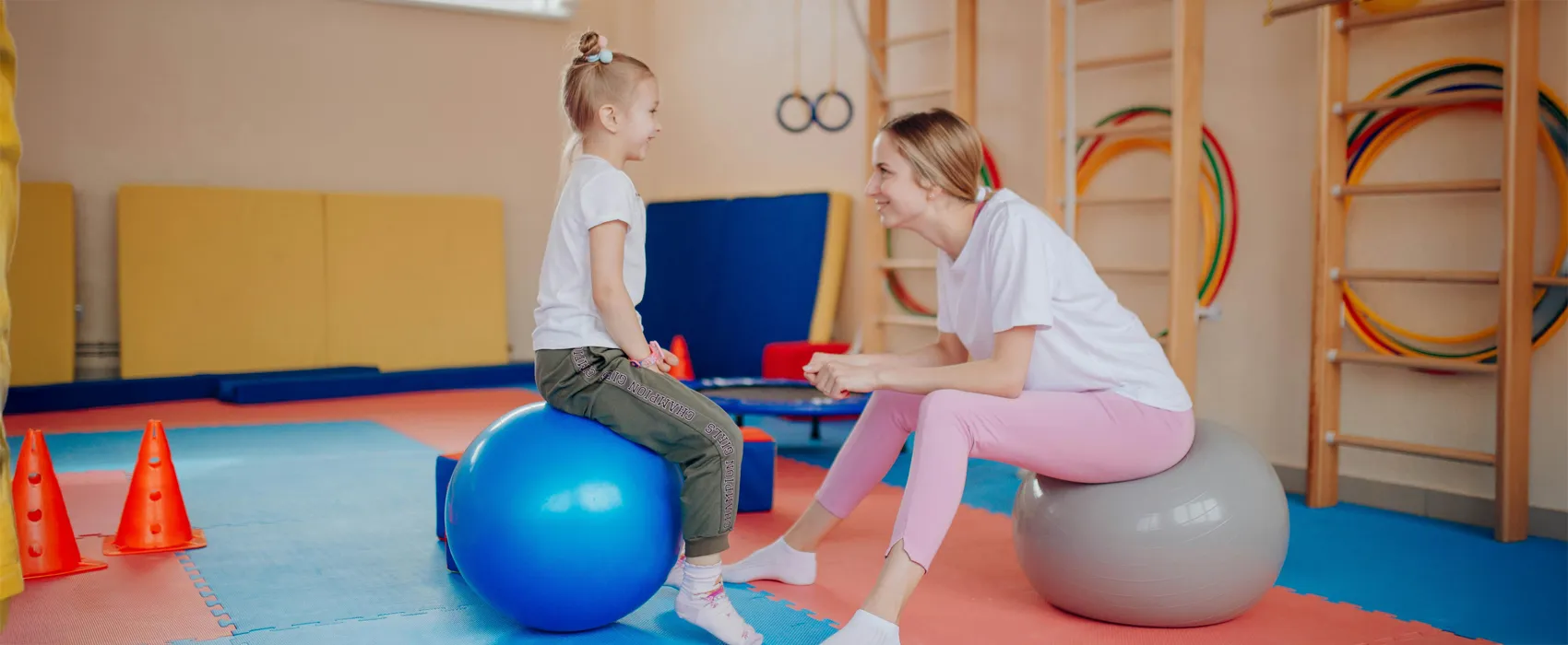 A smiling mother and daughter sitting on fitness balls in a colorful gym setting, sharing a happy moment. Perfect inspiration for motivational gym quotes about family fitness and bonding.