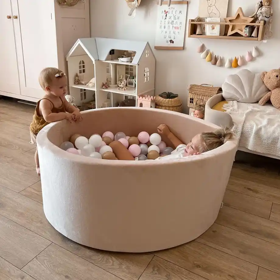 Two toddlers enjoying indoor games in a beautifully designed playroom. One child relaxes in a pastel pink ball pit filled with soft-colored balls, while the other stands nearby, creating a playful and cozy atmosphere perfect for indoor activities.