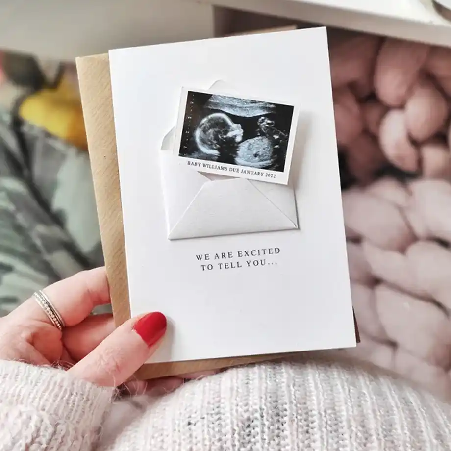 A personalized pregnancy announcement card featuring a sonogram photo in a small envelope with the text "We are excited to tell you..." held by a person with a cozy background.