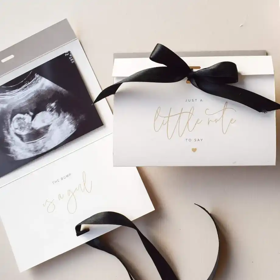 A stylish pregnancy announcement featuring a folded card with a black ribbon, a sonogram photo, and the message "The bump is a girl," offering a sophisticated and heartfelt way to share the news.