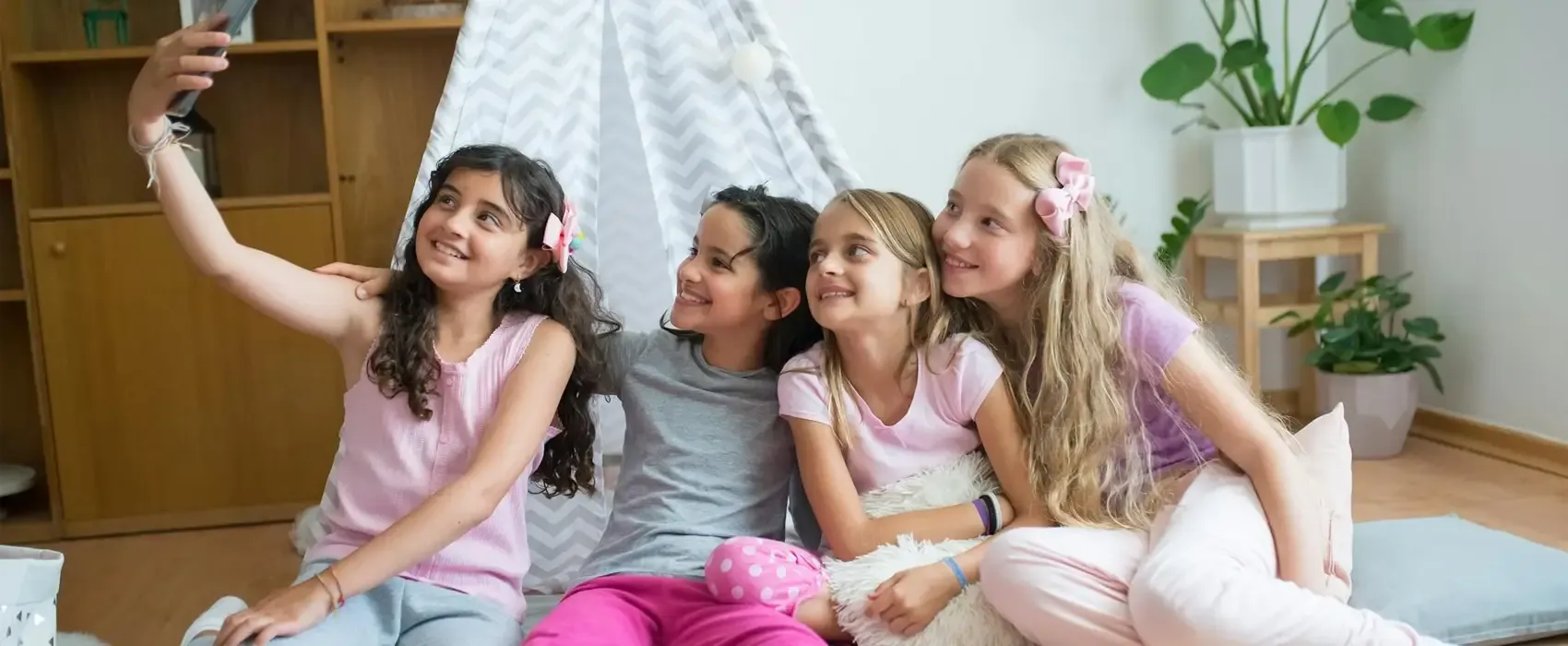 A group of four cheerful girls sitting together on pillows during a sleepover, smiling and taking a selfie with a smartphone. Perfect bonding moment showcasing fun things to do at sleepovers.