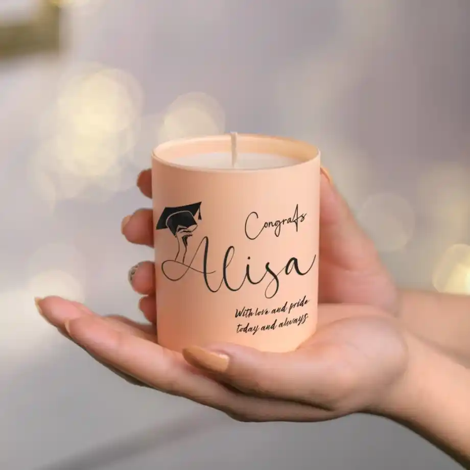 A peach-colored candle held in hands, featuring the text 'Congrats Alisa' with a graduation cap graphic and a heartfelt message, 'With love and pride today and always.' A thoughtful personalized graduation gift for her.