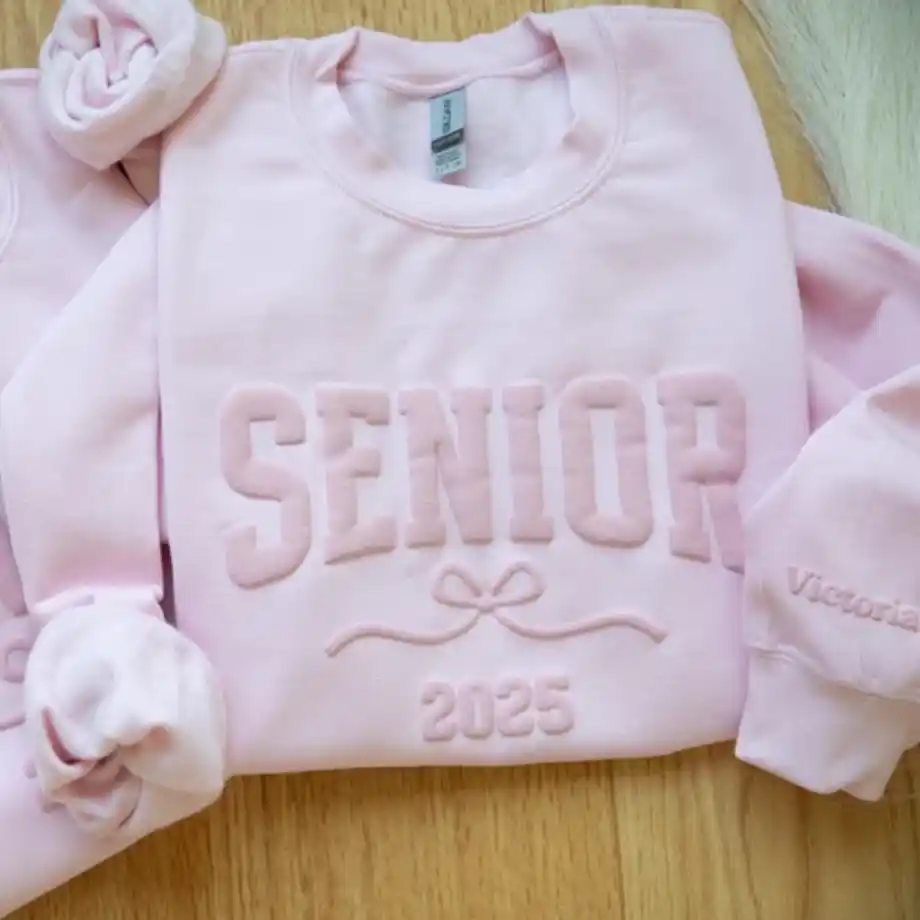 Two pastel pink sweatshirts featuring puff print designs with the text 'SENIOR 2025' and a bow graphic. One sleeve is personalized with the name 'Victoria,' making it a thoughtful gift for a graduating senior from the Class of 2025.