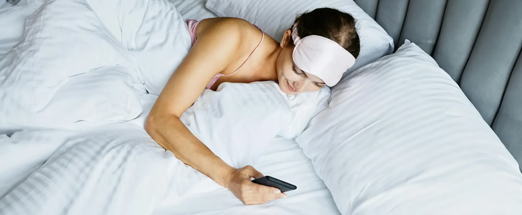 A woman lying in bed with white bedding, wearing a pink sleep mask, and smiling while reading a good morning message for her smartphone.