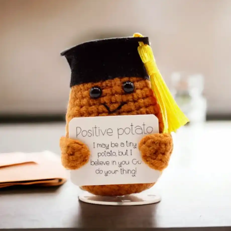 A handmade crocheted potato plush wearing a graduation cap with a yellow tassel, holding a card that reads, 'Positive potato: I may be a tiny potato, but I believe in you. Go do your thing!' in a decorative gift box with shredded paper lining.