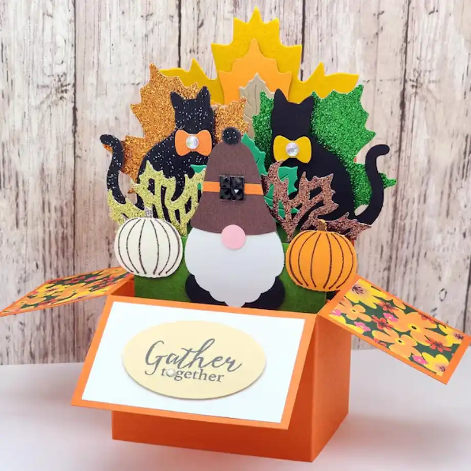 A vibrant pop-up card with a gnome, two black cats adorned with orange bows, glittery autumn leaves, and pumpkins. The card displays "Gather together" on the front, ideal for conveying warm and happy thanksgiving messages.