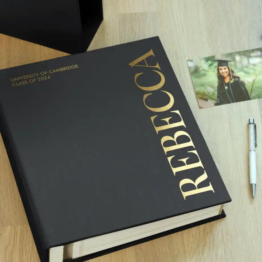 A black hardcover graduation scrapbook personalized with gold text reading 'Rebecca,' featuring the University of Cambridge Class of 2024, displayed alongside a graduation photo and a pen on a wooden surface.