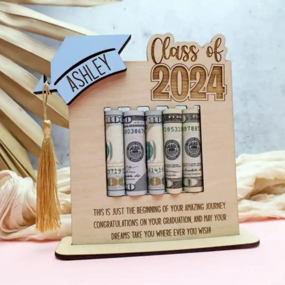 A wooden graduation gift featuring a money holder with rolled dollar bills, a decorative graduation cap labeled 'Ashley,' and the text 'Class of 2024.' Below, a heartfelt message congratulates the graduate and wishes them success on their journey.