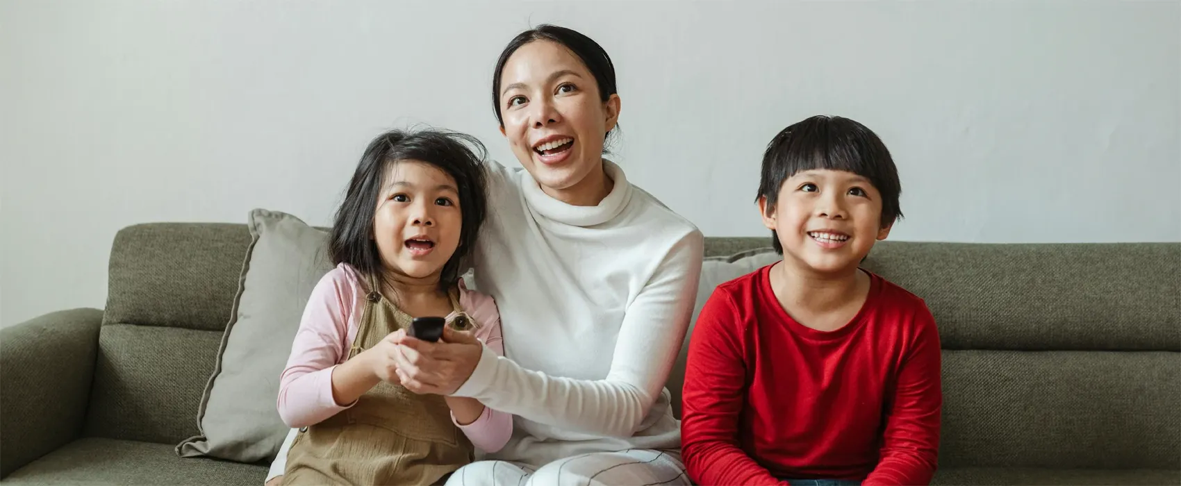 A cheerful mom sits on the couch with her two kids, watching a movie together. The young girl holds the remote control, and everyone is smiling. Perfect family bonding moment while enjoying the best animated movies.