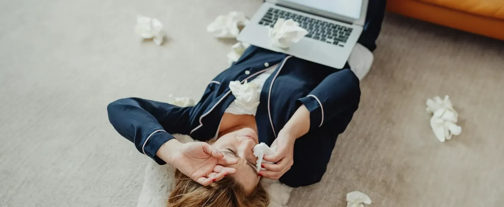 A tired woman lying on the floor in pajamas surrounded by tissues and a laptop, symbolizing the use of bulletproof excuses to get out of work.