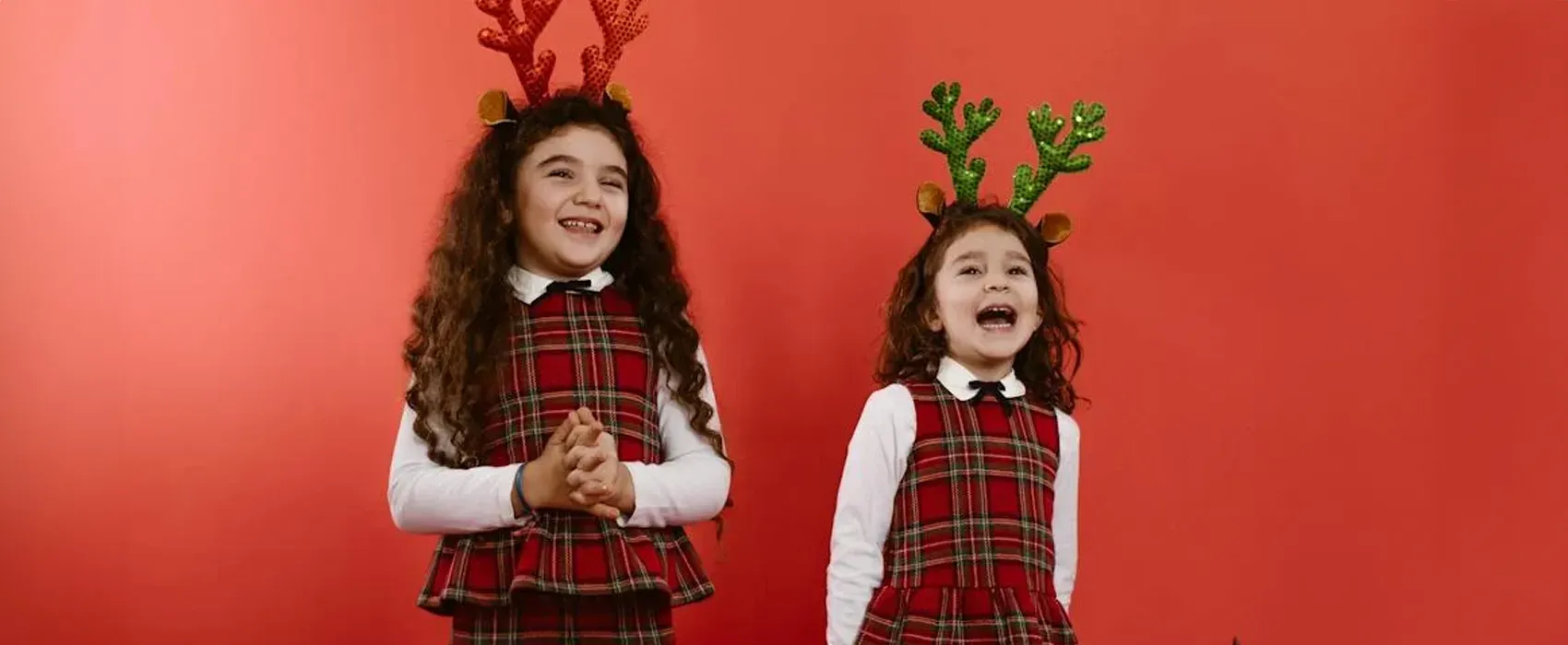 Two young girls wearing festive plaid dresses and reindeer antlers are laughing joyfully against a bright red background, creating a cheerful holiday vibe perfect for sharing Christmas jokes.