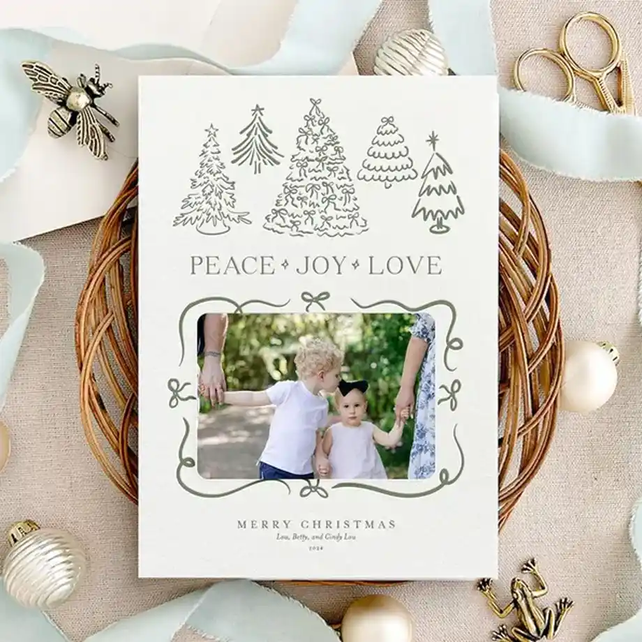 A beautifully designed Christmas card featuring hand-drawn trees, the words "Peace • Joy • Love," and a family photo surrounded by festive decorations, including ornaments, ribbon, and scissors. A perfect way to send heartfelt Christmas wishes.