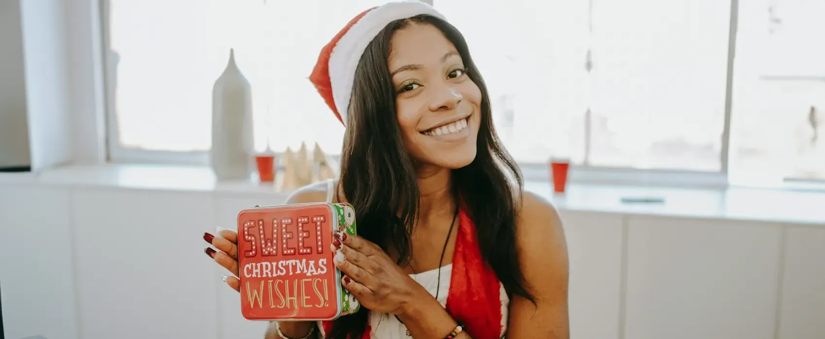 Smiling woman wearing a Santa hat and red scarf, holding a festive gift box with the words "Sweet Christmas Wishes" printed on it, in a brightly lit room.