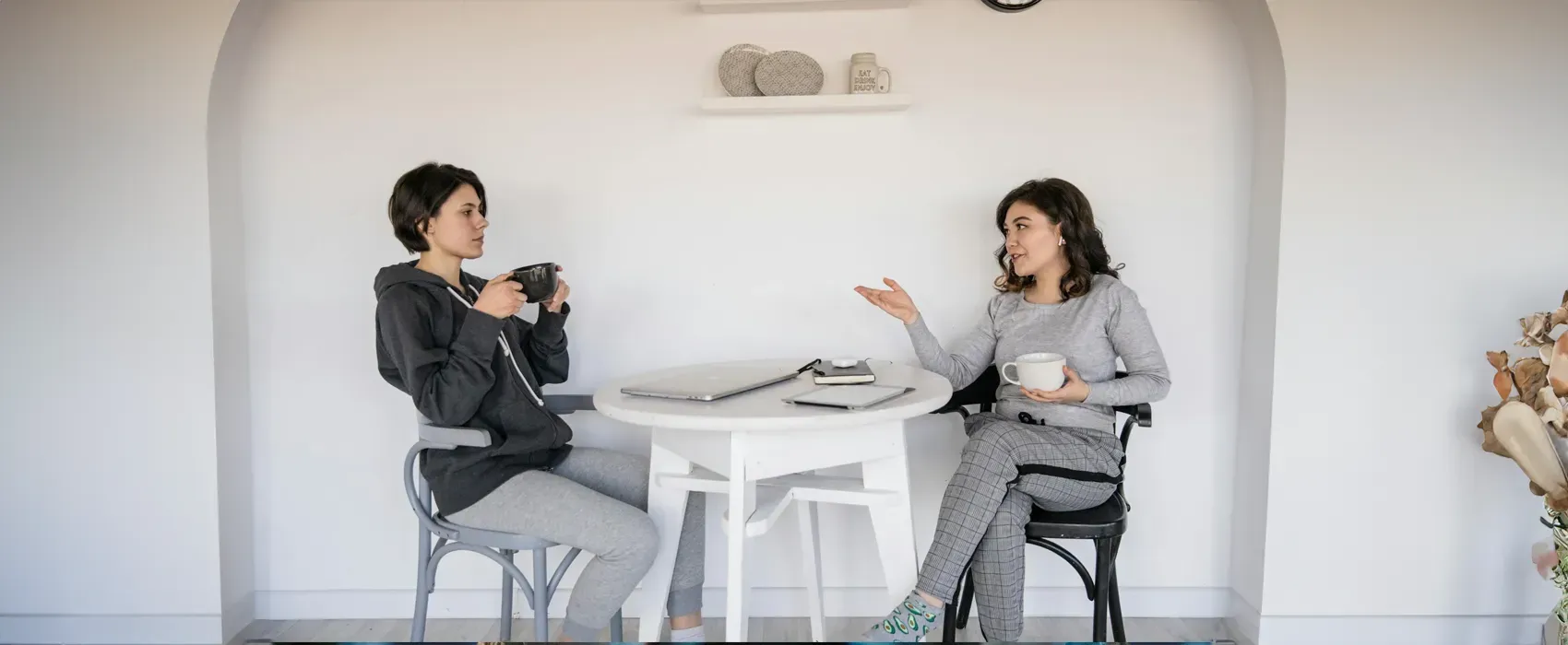 Two women sitting at a small round table with coffee mugs in hand, engaged in a friendly and relaxed conversation. A notebook and laptop are on the table, creating a cozy setting ideal for exploring conversation starters.