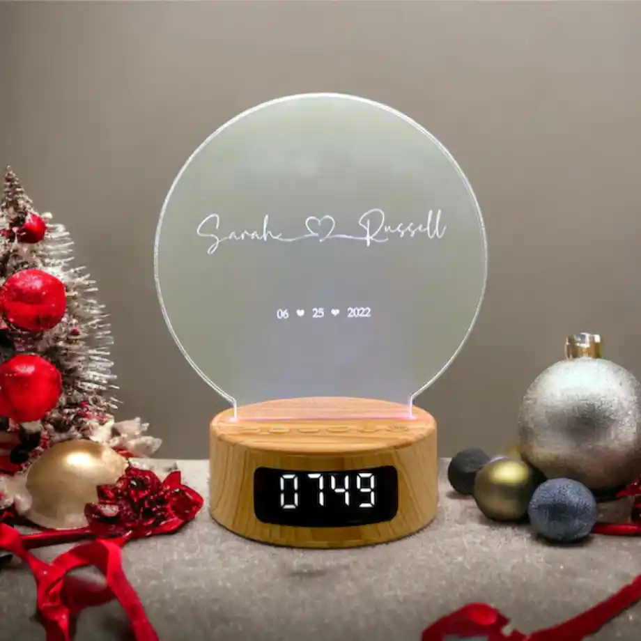 A custom LED acrylic clock featuring the names Sarah and Russell with a heart symbol, the date 06.25.2022, and a wooden base with a digital display, surrounded by Christmas-themed decorations.