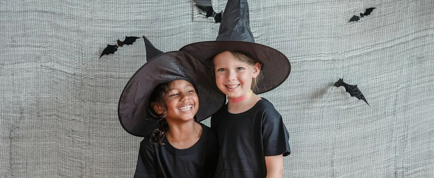 Two cheerful children dressed as witches, standing in front of a Halloween-themed backdrop with bats, perfect for sharing Halloween jokes and festive fun.