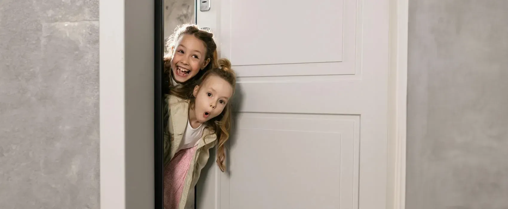 Two cheerful kids peeking from behind a white door, laughing and looking surprised, perfect for a fun moment with knock knock jokes.