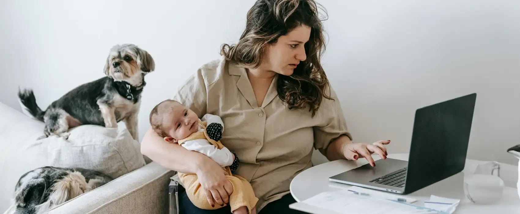 A mother holding her baby while working on a laptop, accompanied by two small dogs on a couch, portraying a relatable scene of drafting a maternity leave out of office message while managing work and home responsibilities.