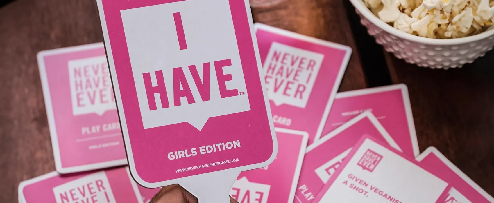 A deck of pink "Never Have I Ever" cards, including an "I Have" paddle from the Girls Edition, placed on a wooden table alongside a bowl of popcorn, perfect for game night fun.